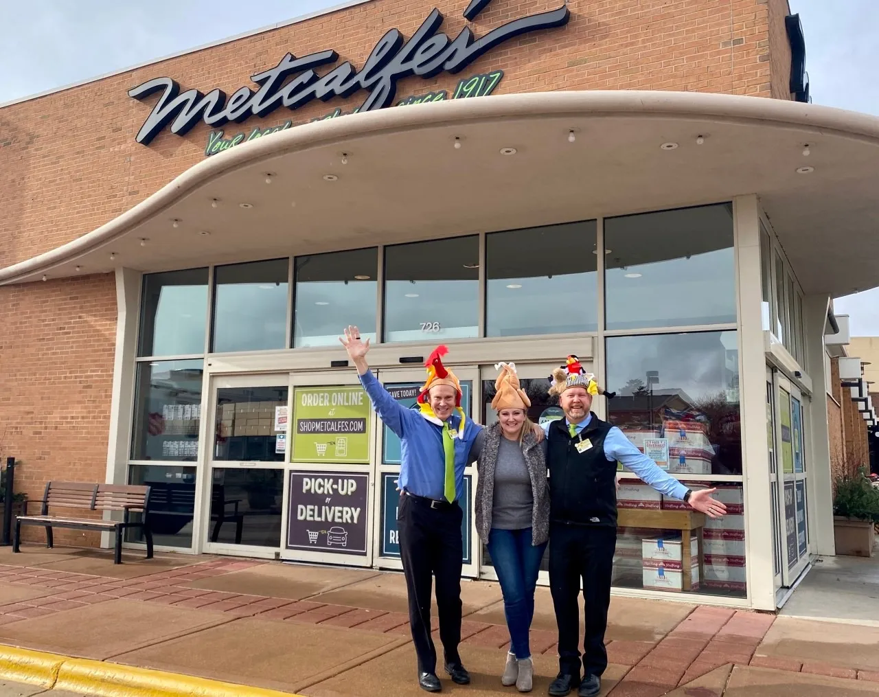 Three people wearing hats, including turkey ones, outside of a Metcalfe's store