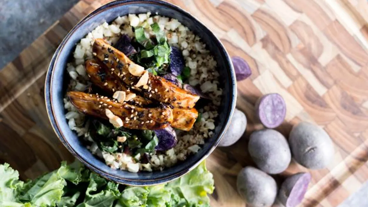 A bowl with two strips of chicken atop cooked grains sits on a wooden cutting board, which is on a gray marble surface. Lettuce and cut purple potatoes are next to it.