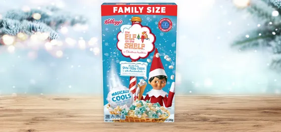 A box of Elf on the Shelf cereal sits on a wooden counter with snow covered lighted pine trees on either side.