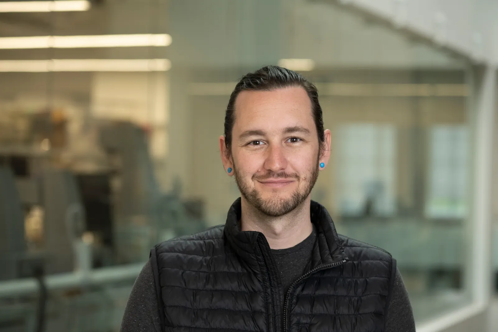 Chris Bryson, in a black shirt and puffer vest, stands in front of a glass-walled lab area.