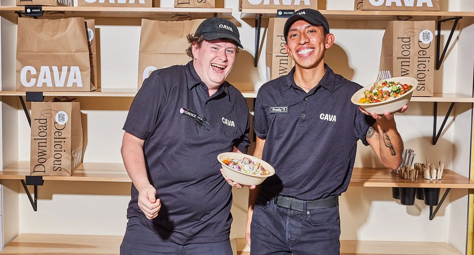 Two smiling men wearing black T-shirts and black ballcaps with Cava branding. One man is holding up a bowl from Cava.