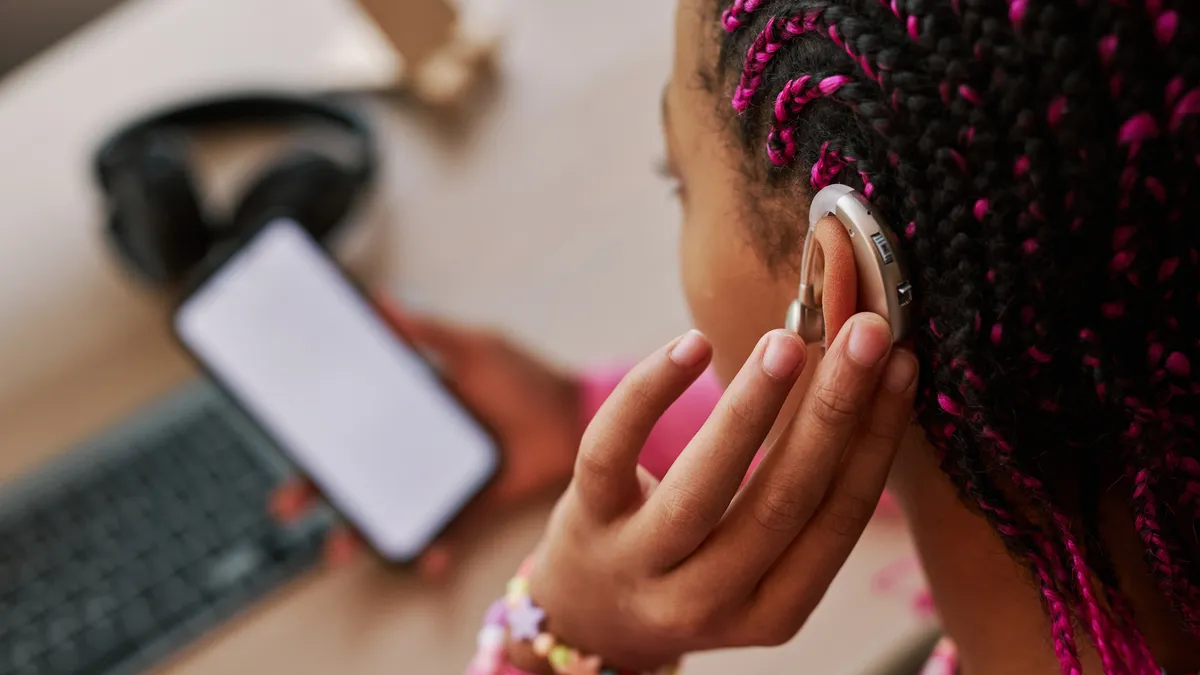 A student, whose shown on the side of their head, has a hearing device in their ear. There is a desk with a laptop in front of the person and a cellphone in their hand.