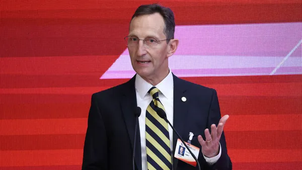 A man in a suit and tie speaks into a microphone in front of a red background.