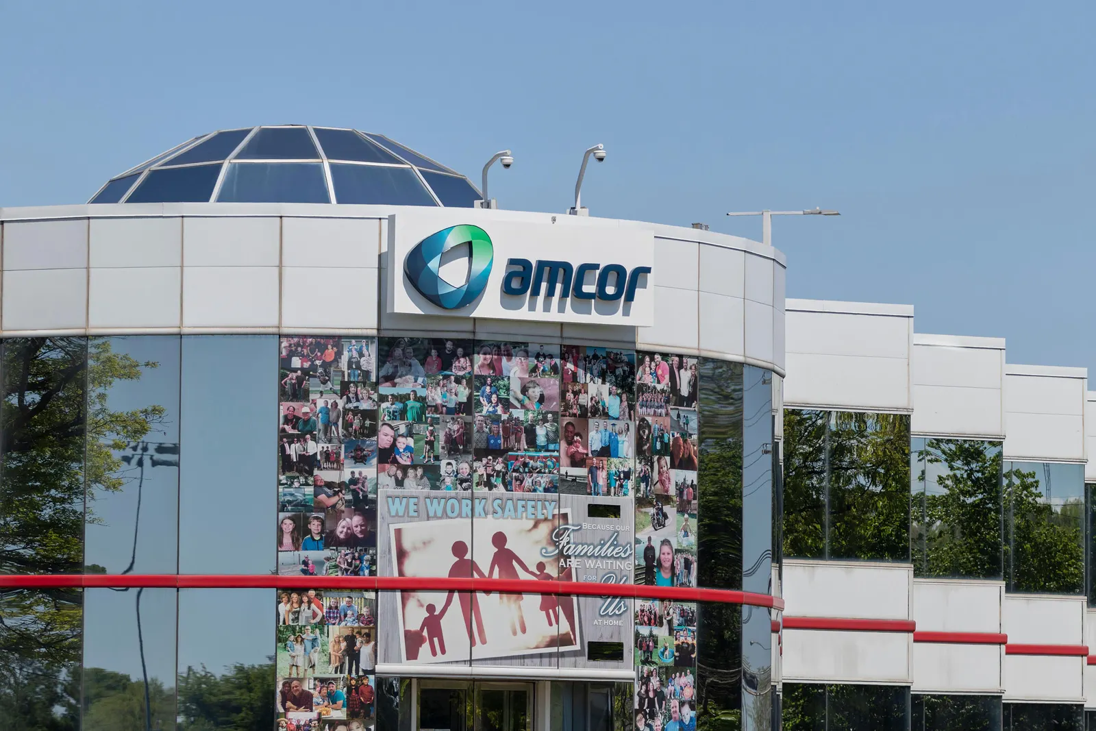 A modern white commercial building with reflective glass windows features a prominent "Amcor" sign at the top. The front of the building displays a collage of employee and family photos, along with a large safety sign that reads, "We work safely because our families are waiting for us at home." 