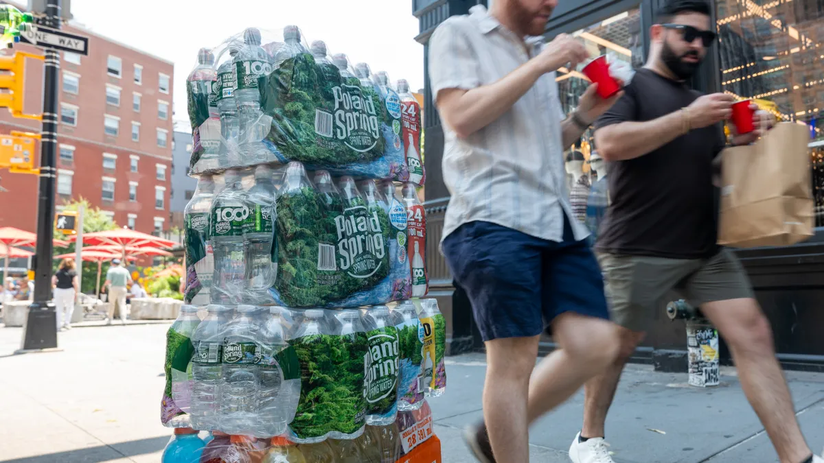 Cases of Poland Spring bottled water sit stacked on a city sidewalk as people walk past.