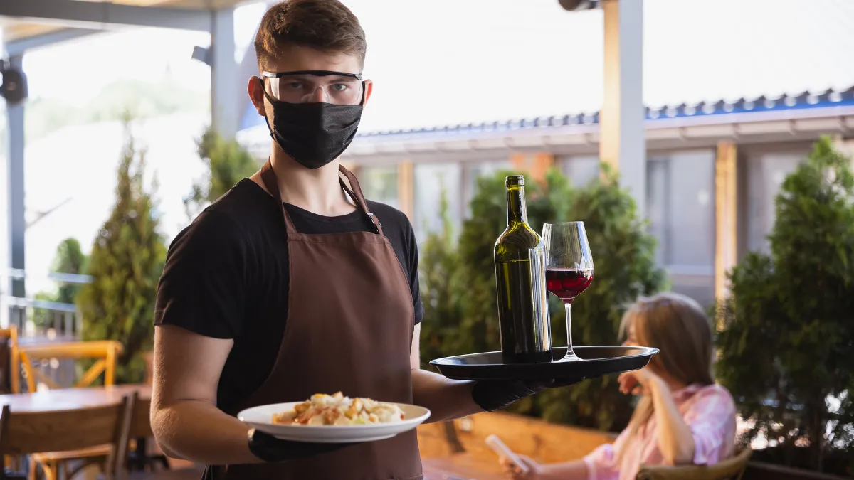 The waiter works in a restaurant in a medical mask, gloves during coronavirus pandemic.