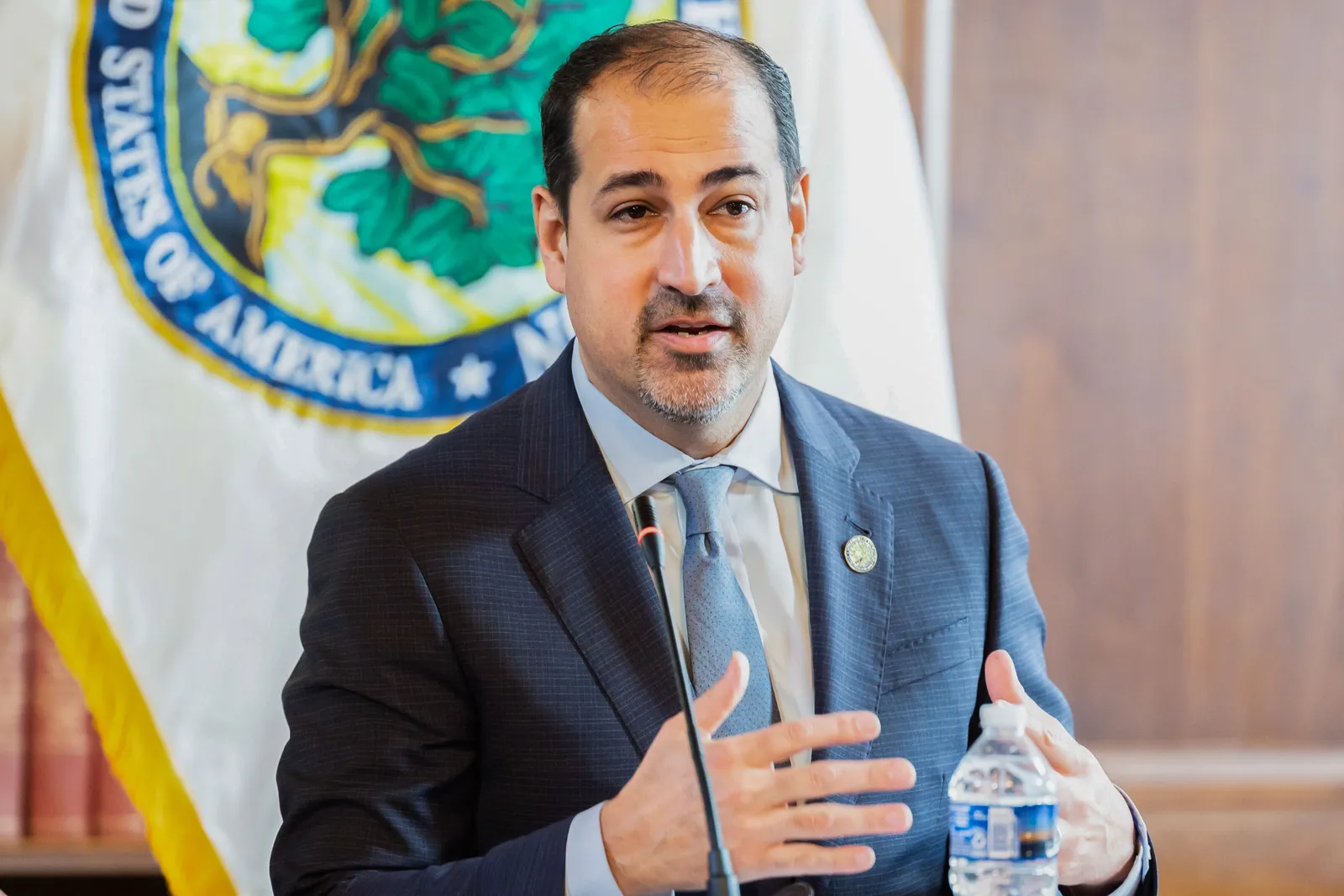 A person wearing a jacket and tie is standing in front of a microphone. There is a water bottle near the microphone. A flag is in the background.