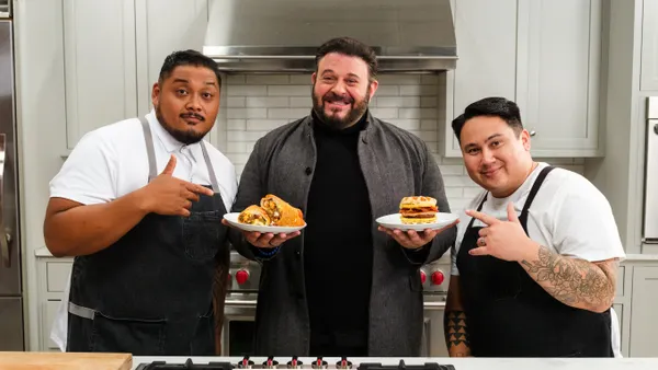 A photo of three people in a kitchen.The one in the middle is holding two plates of food while the other two are pointing at the food.