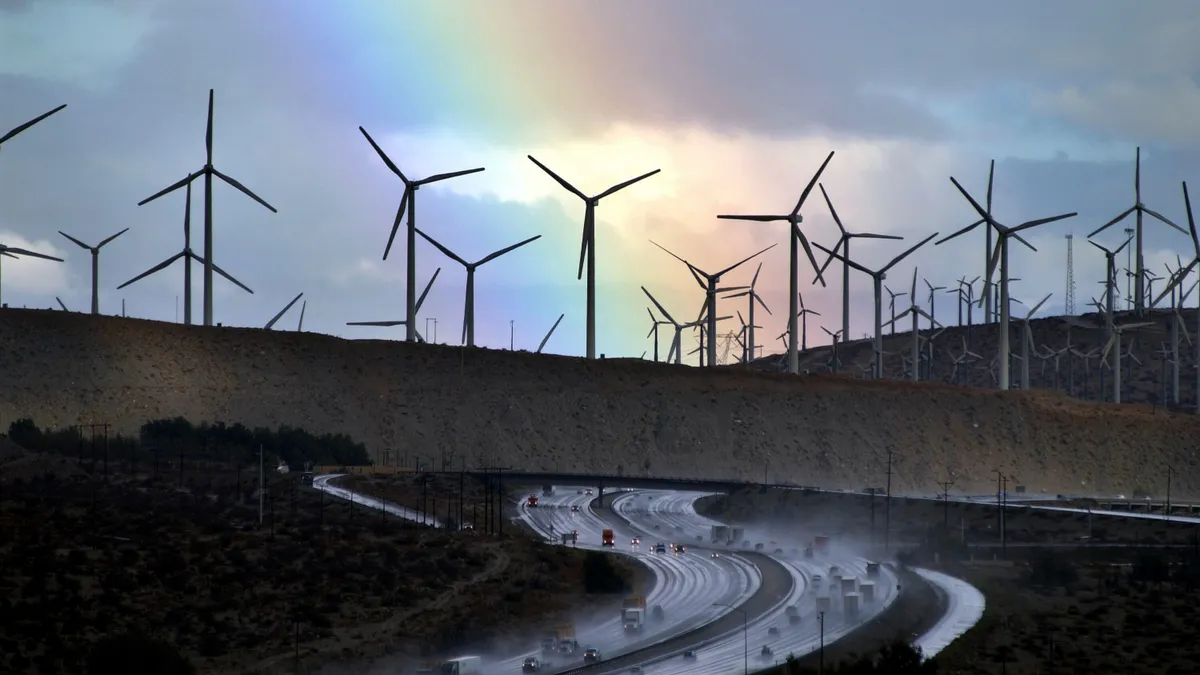 Cars drive on a rainy road while wind turbines are shown behind them in front of a rainbow.