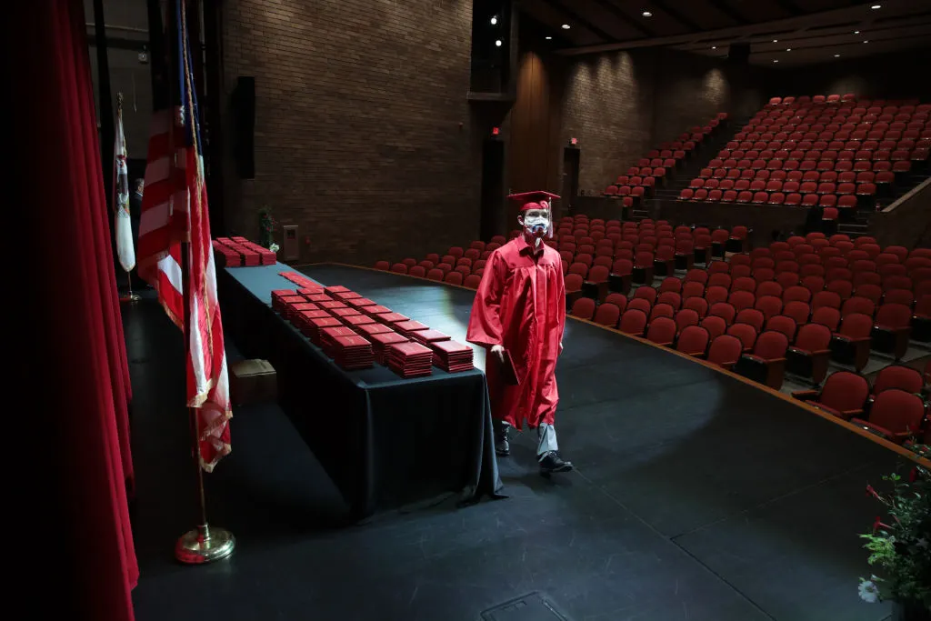 A person in a graduation cap and gown walks across a stage in an auditorium. A table of diplomas is on the stage. No one else in there.