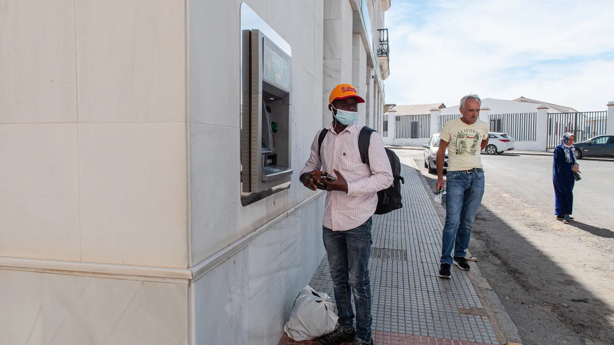 Person stands outside building in front of ATM with things in his hands and plastic bag at his feet, with two people walking by behind.