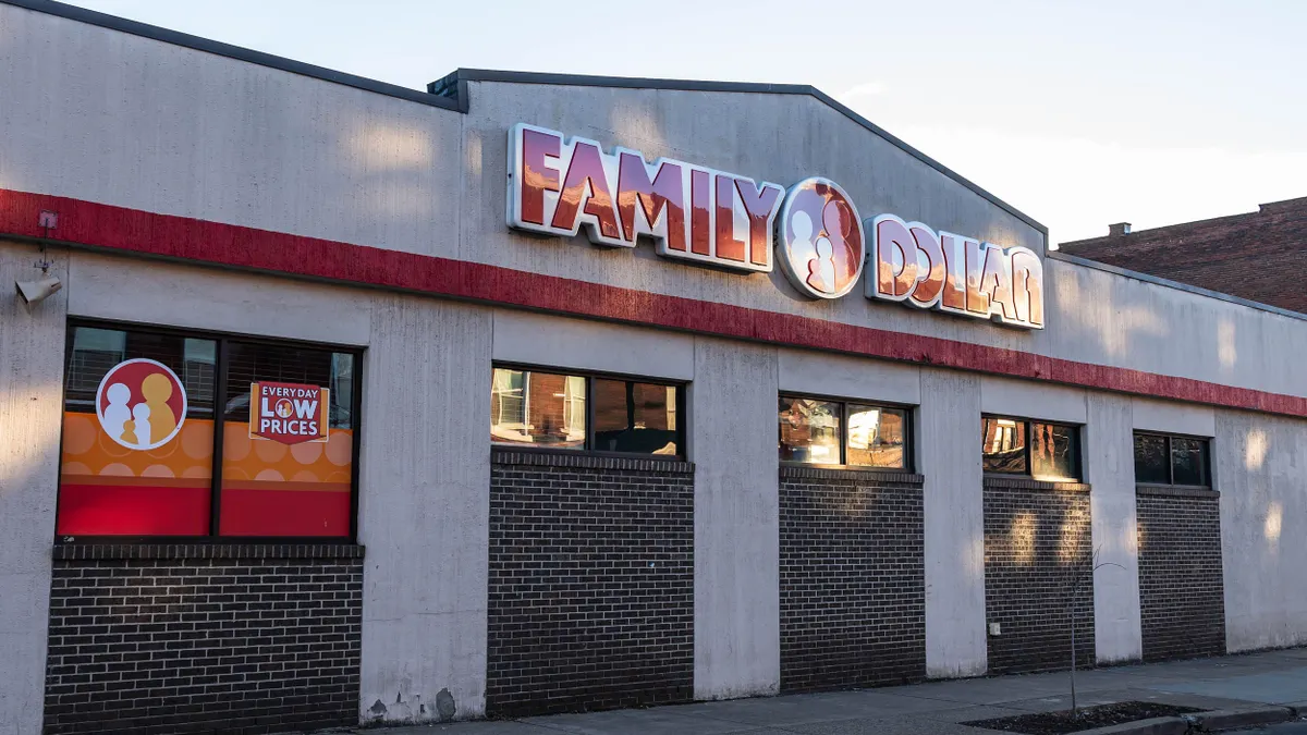 Street-level view of a Family Dollar store building with a red and white sign that reads “FAMILY DOLLAR.” The storefront has several windows with red and orange branding. The wall is a mix of concrete and dark brick, and the sidewalk is visible in the foreground.