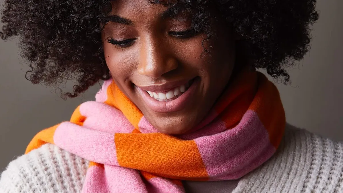 A person wears a striped multi-colored scarf and a white sweater and smiles while looking down.