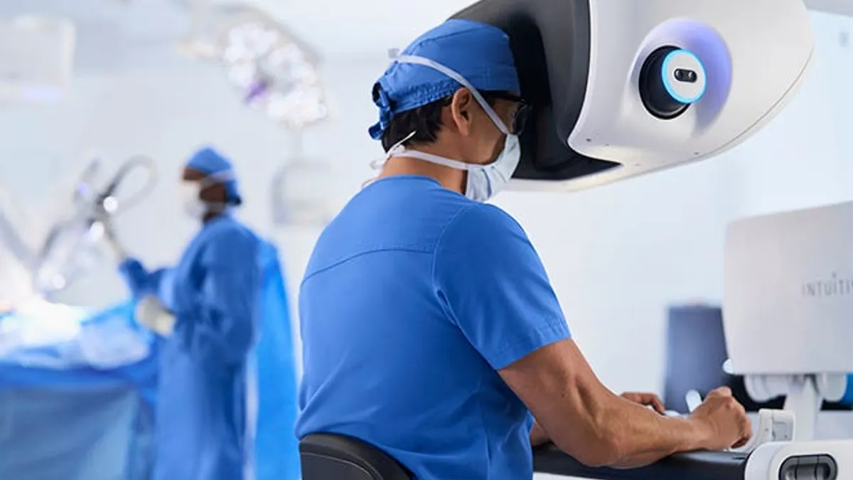 A surgeon sits at the console of a da Vinci 5 robot while another medical professional works in the background.
