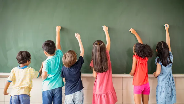 A handful of students are facing a green chalkboard in a classroom. They have their hands raised ready to write on the chalkboard
