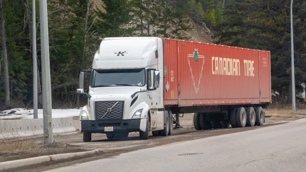 A white Volvo semi-truck with a red Canadian Tire trailer in British Columbia with a wooded hillside in the background.
