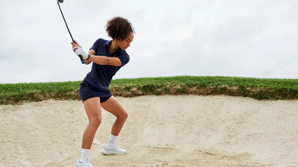 A golfer swings a club and wears a black skort outfit.