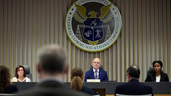 Three people sit at a long table in front of a crowd and under a sign that says "United States Federal Communications Commission."