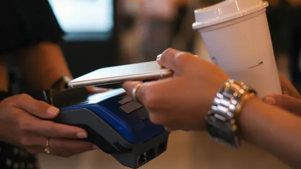 Close-up shot of unrecognizable female hands holding her smartphone above card reader while paying contactless for her takeaway coffee held in other hand