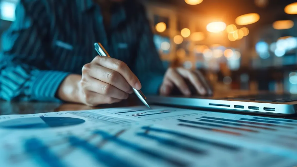 Person's hand writing with a pen next to a laptop.