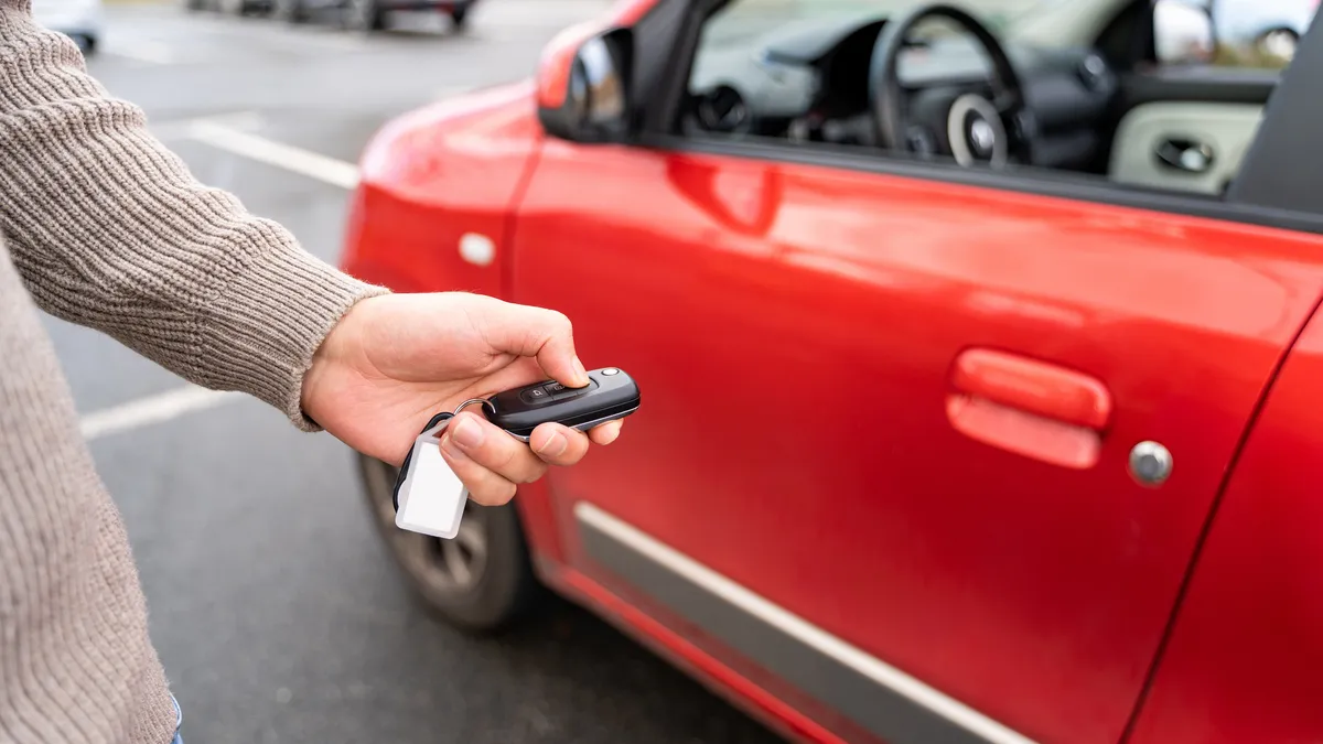 Hand holding electronic remote key pushing button near red rental car