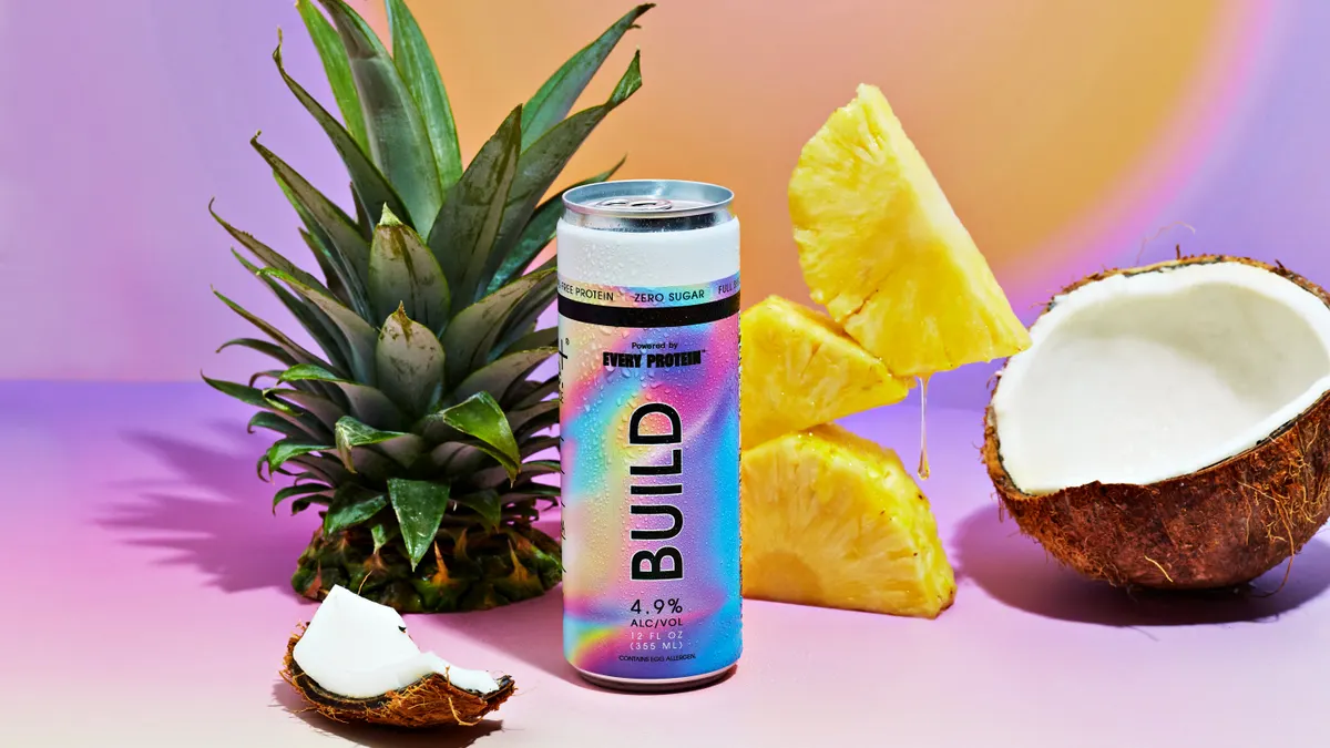 A rainbow swirled can of Build next to a pineapple top and slices and pieces of a cracked coconut on a pastel background.