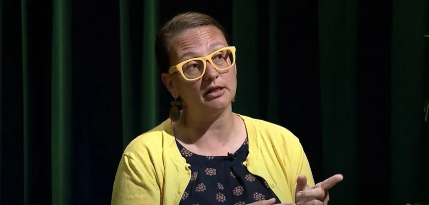 A person wearing yellow glasses, large earrings and a yellow cardigan speaks.