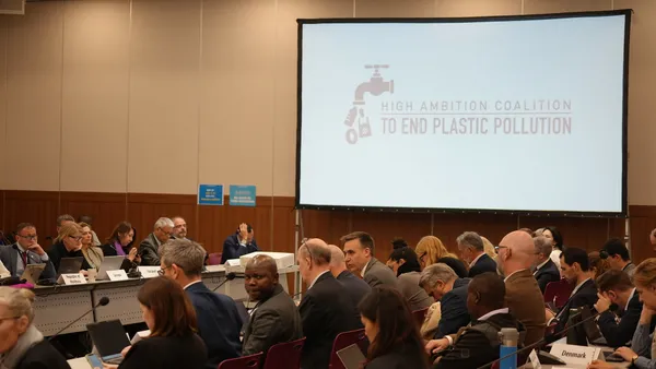 A room of people sit in chairs at a meeting, with a screen saying "High Ambition Coalition to End Plastic Pollution" in the background