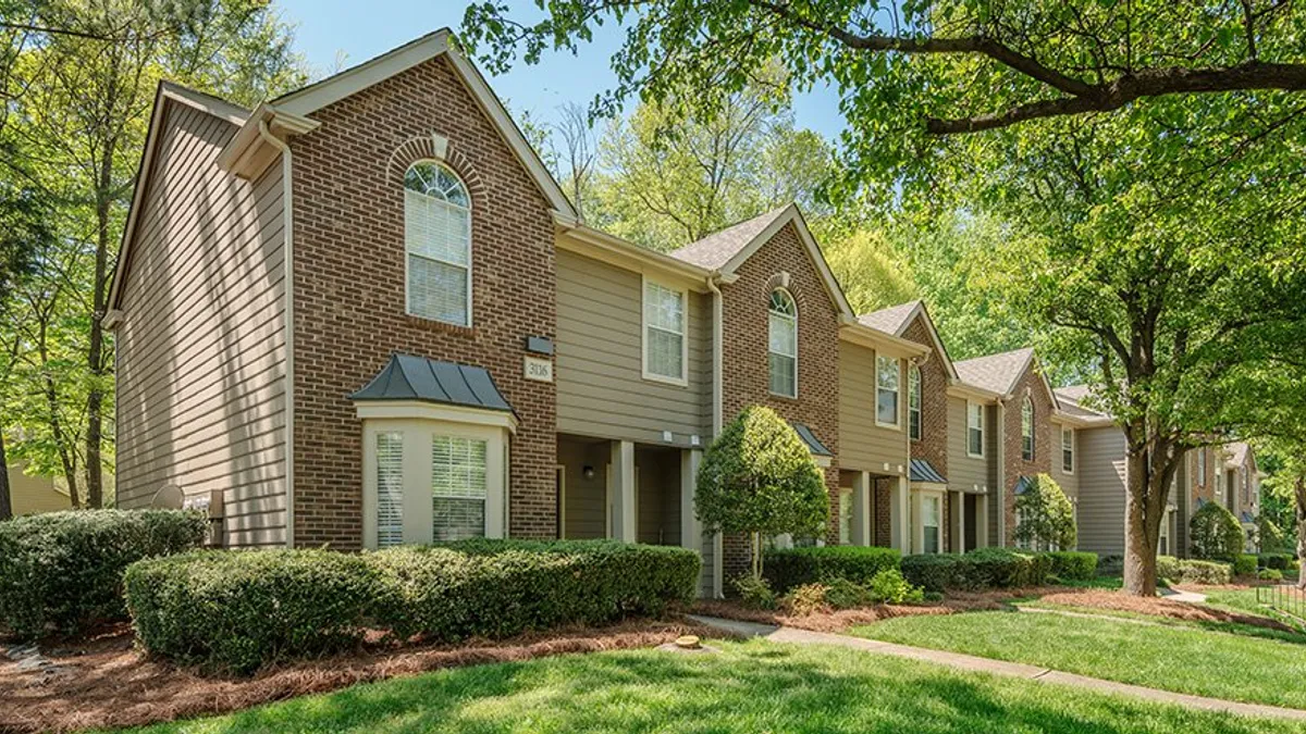 Two-story brown, brick apartments with grass and tree in the foreground.