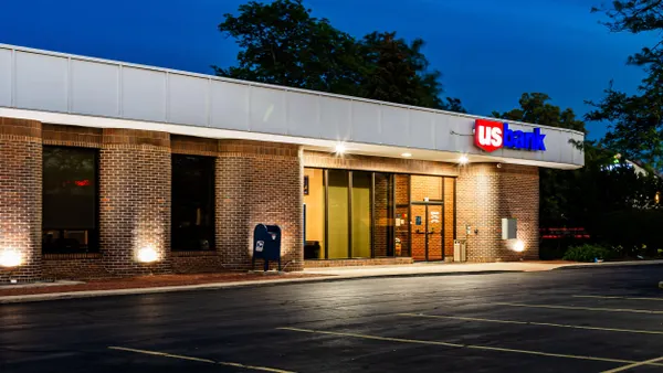 A brick US Bank branch captured at dusk with illuminated signage and lights around the building. The parking lot in front is empty, and there’s a blue USPS mailbox near the entrance.