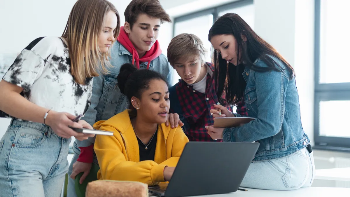 High school students sitting together at desk and using laptop and talking during break in classroom