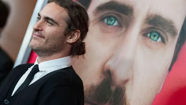 Actor Joaquin Phoenix stands smiling in front of his own face on the poster for the film "Her."