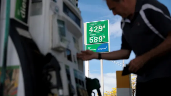 A person at a fuel pump with a green sign showing gas prices in the background.