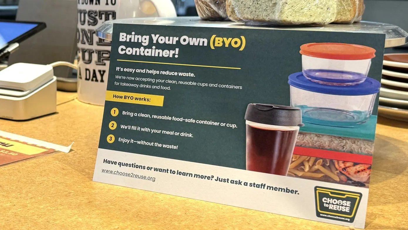 A person's hand holds a reusable coffee cup with a "Choose to Reuse" sticker on the side, near a foodservice establishment sign explaining the bring your own container initiative.
