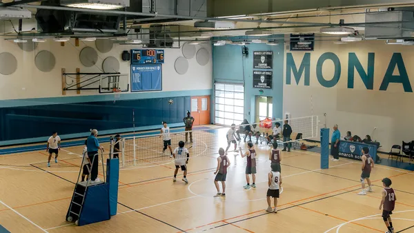 A wide angle photo of a school gym shows a volleyball game with several players on each side of the net.