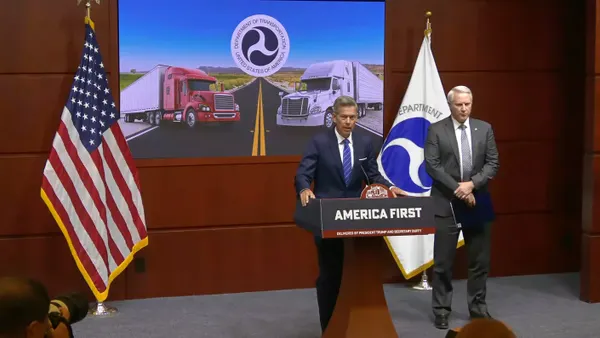 Two people stand at a podium with a sign that says, "America First." Behind them, a screen projects an image of two semi-trucks displaying the Department of Transportation logo.