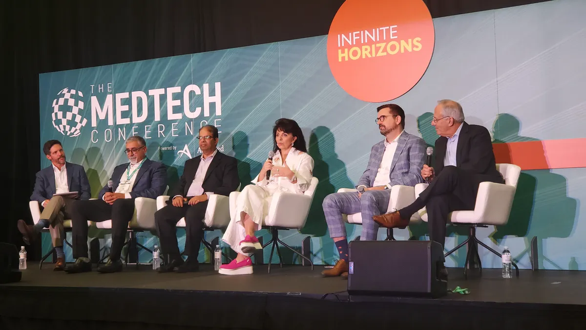 Six people sit on a stage in front of a sign that says the MedTech Conference. A person in the center holds a microphone.