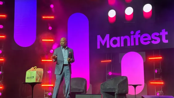 A man speaks on a stage with a Dollar General bag in front of him. The word "Manifest" is displayed behind him.