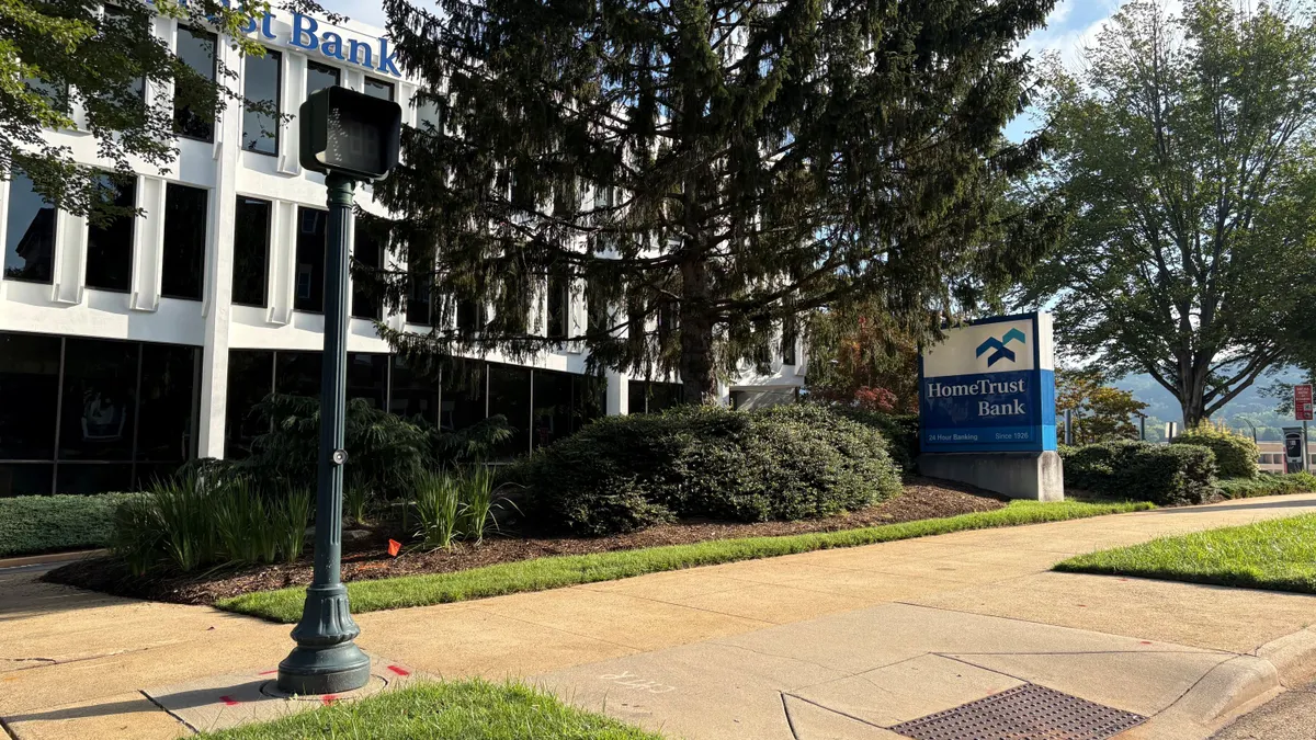 A sign in front of a white and black office building and next to an evergreen tree reads "HomeTrust Bank."