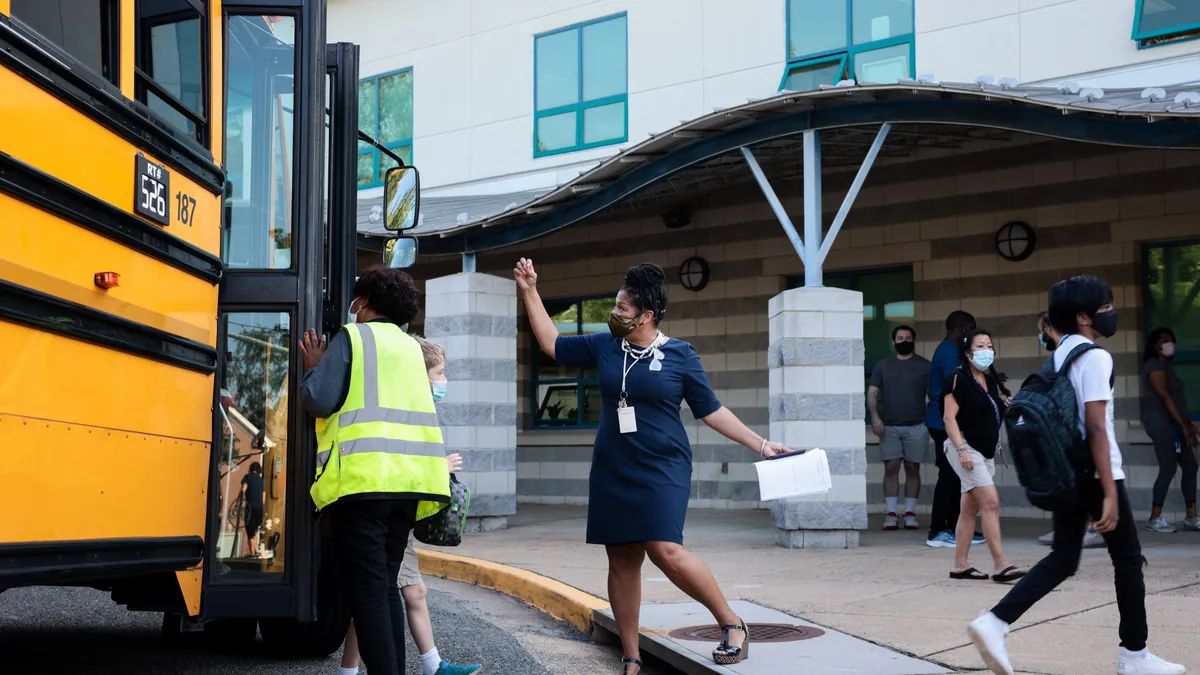 An adult stands outside near a yellow school bus parked alongside the curb near a school. Students are walking off the bus and toward the school.