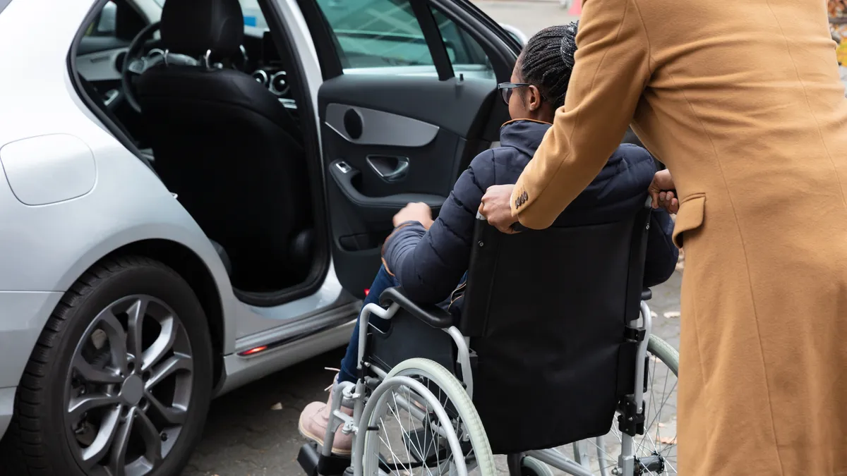 A person in a wheelchair is being pushed by another person toward the open rear door of a sedan.