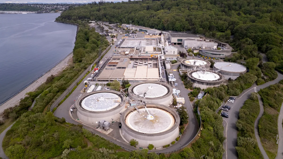 Aerial view of a waste treatment plant, with massive cylindrical holding tanks, surrounded by trees and water on the left.