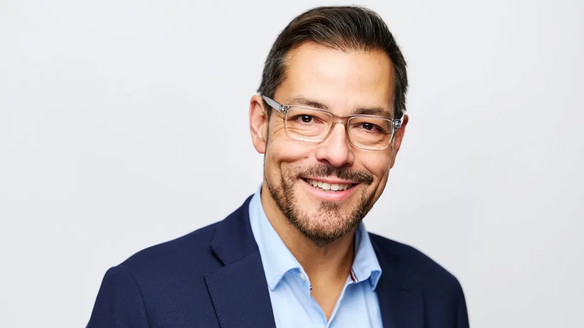 A smiling man with a beard in a blue shirt and blue jacket with lapels.
