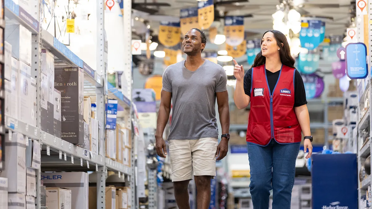 A Lowe's employee in a red vest walks alongside a customer through a store aisle.