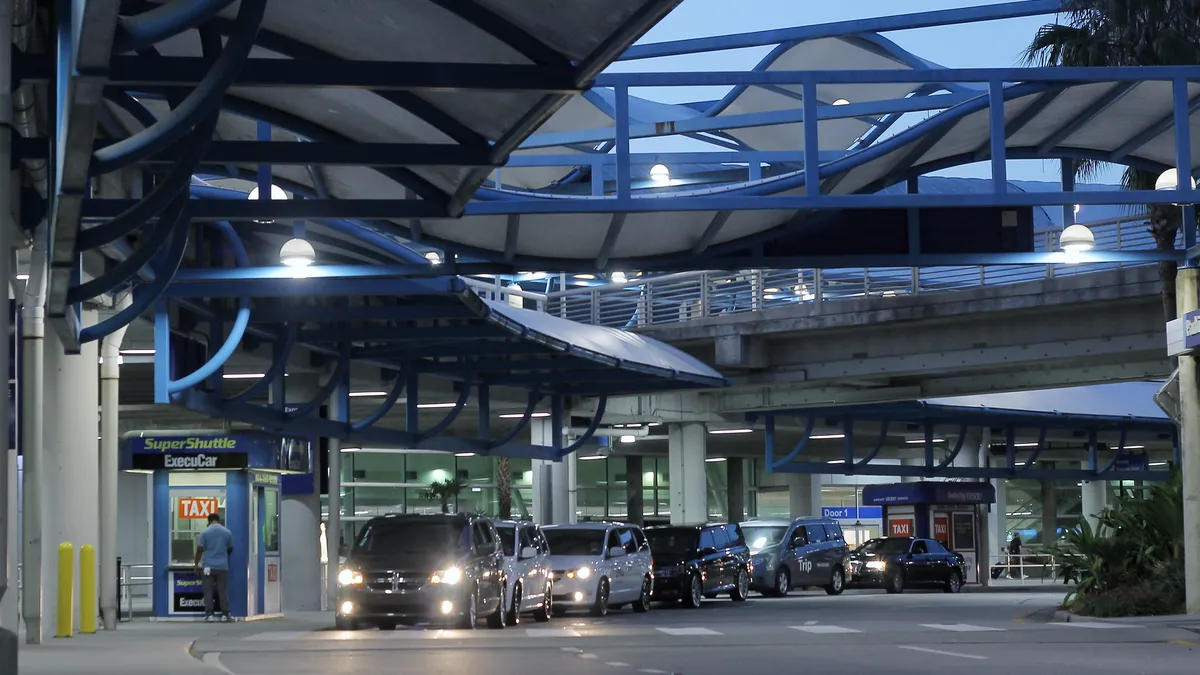 Cars, with their headlights on, drive under a canopy at an airport terminal in the wet night. The fronds of palm trees are visible in the background.