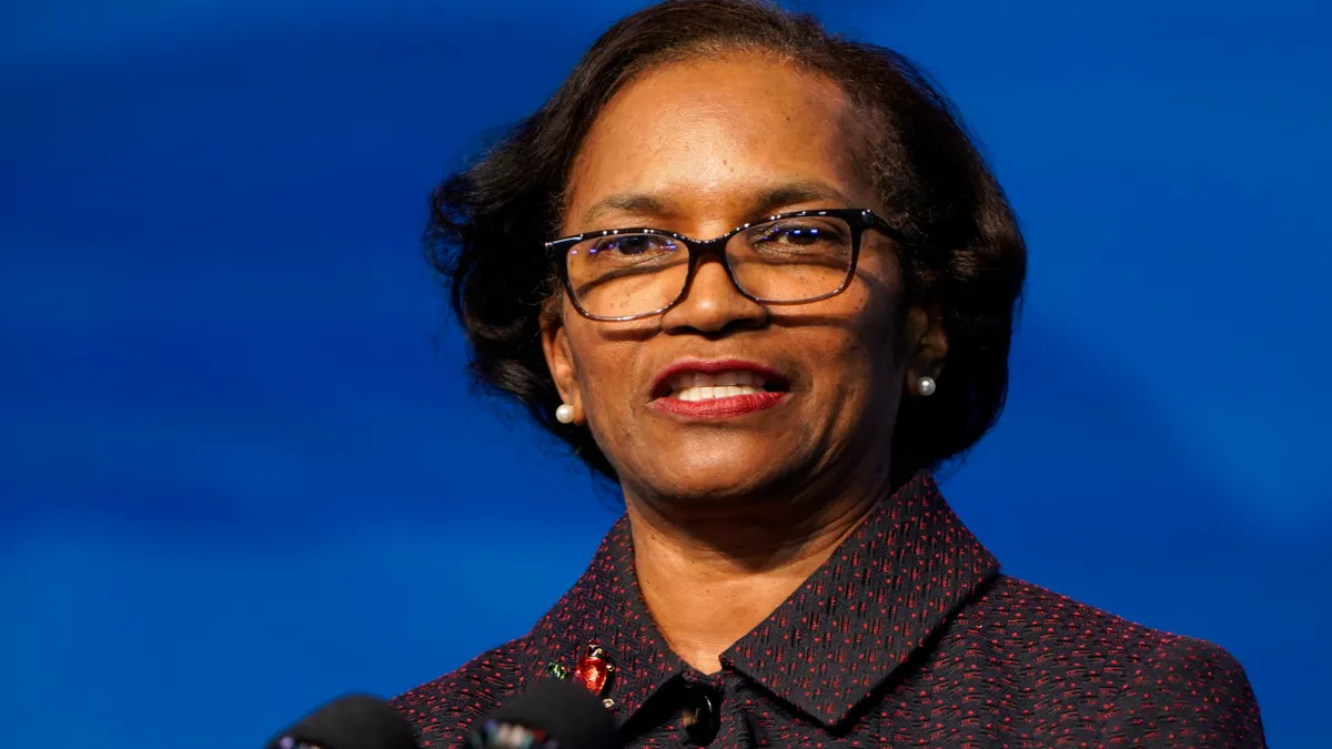 A person wearing glasses speaks into microphones in front of a solid blue background.