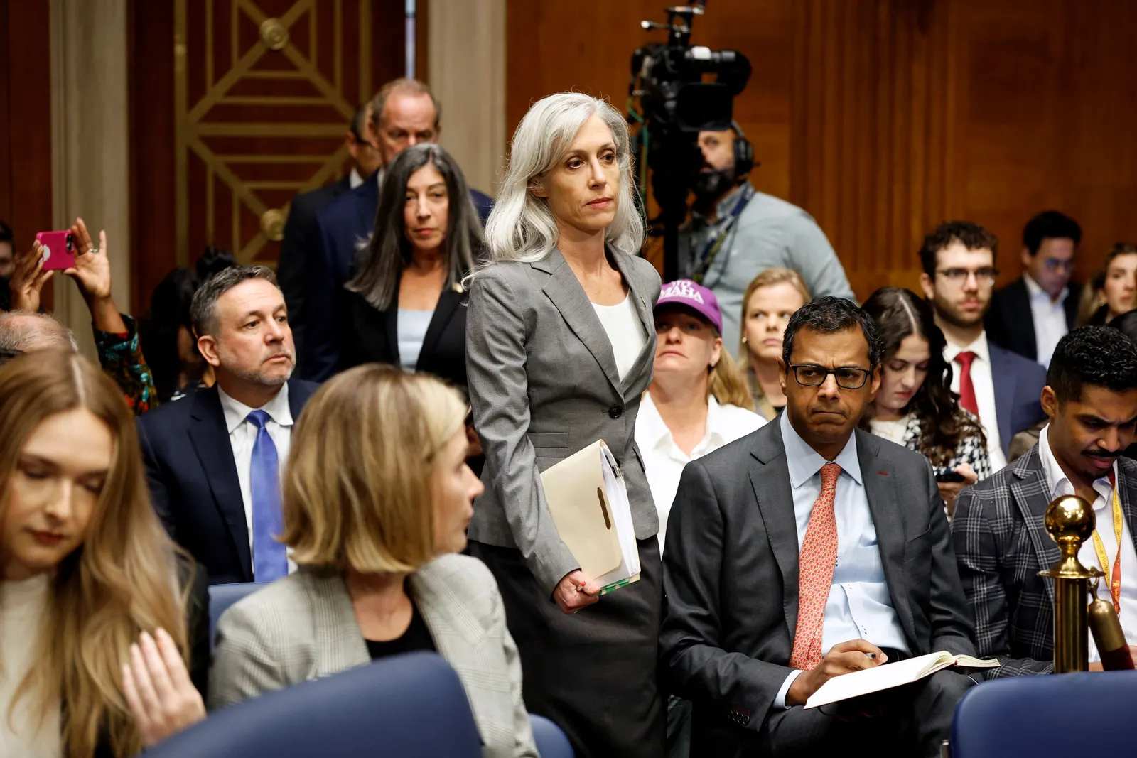 A woman in a blazer stands holding a folder in the middle of various people in business attire sitting closely around her. A standing camera with a man operating it and other people stand in the background.