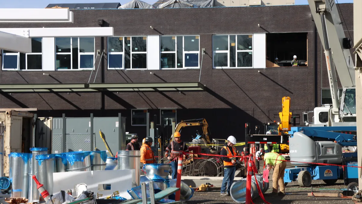 A photo of construction workers walking around a jobsite at a tall, stone building during a bright, blue-skyed day. There are lines of tape separating walkways.