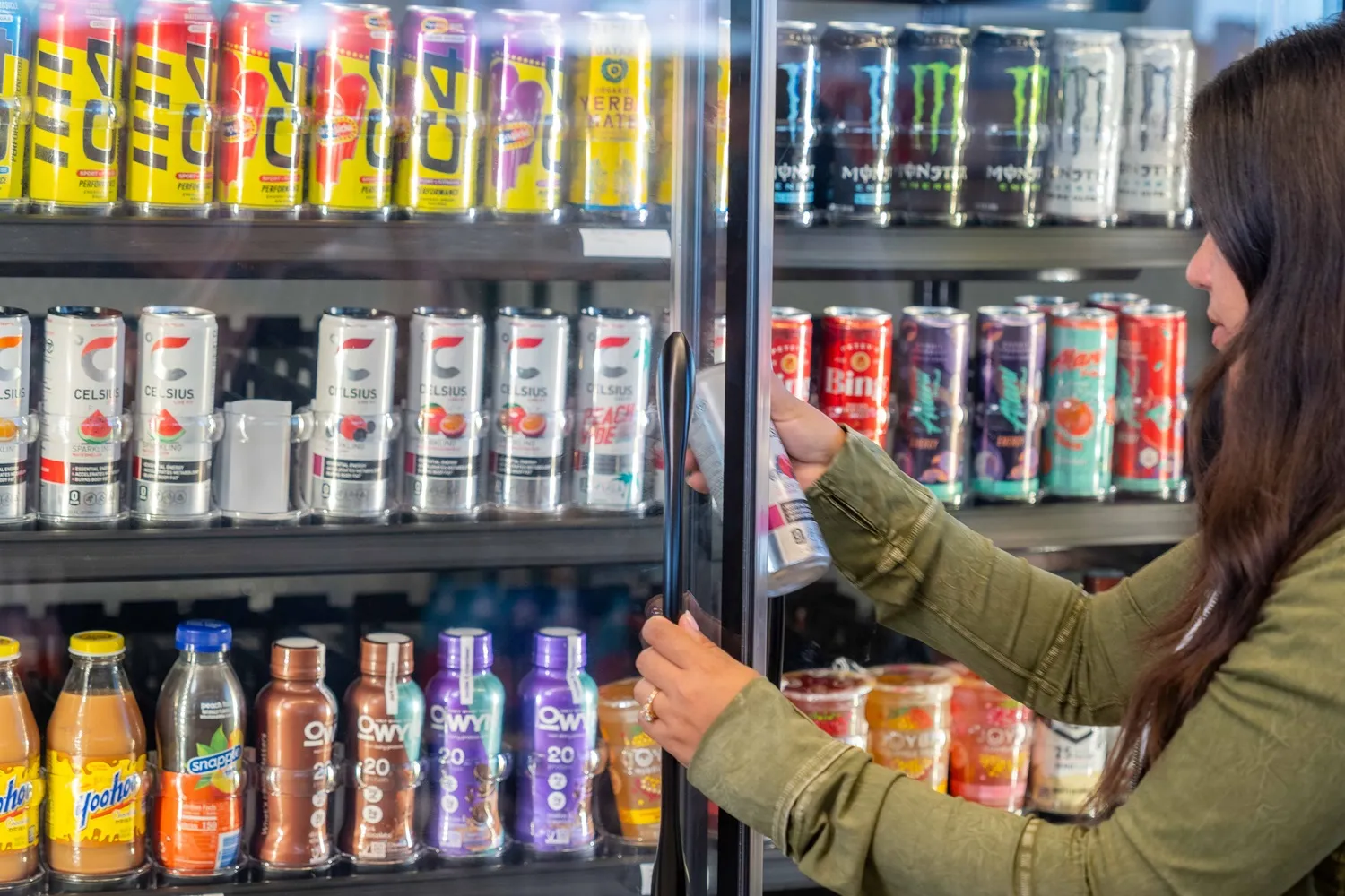 A photo of a number of beverages in a cooler case inside a store. some of the cans say Celsius, Monster and C4.