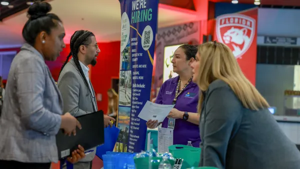 Individuals speak across a table with a "we're hiring" sign in the background.